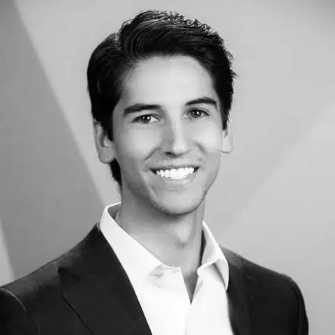 Young man with dark hair wearing a suit jacket and white shirt, smiling at the camera in a studio setting.