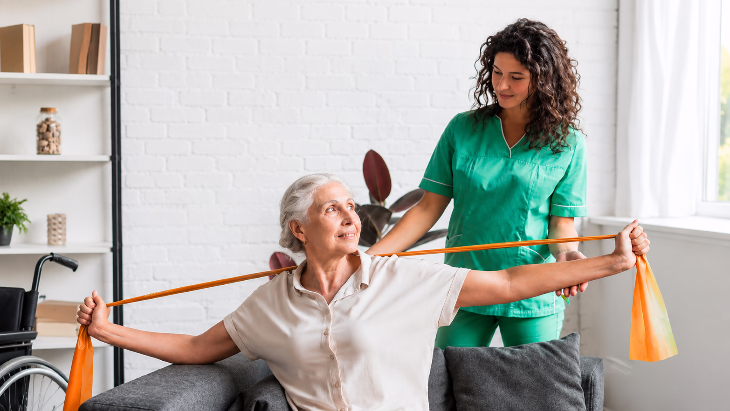 Elderly woman stretching arms with an orange band while a caregiver assists her in a bright room.