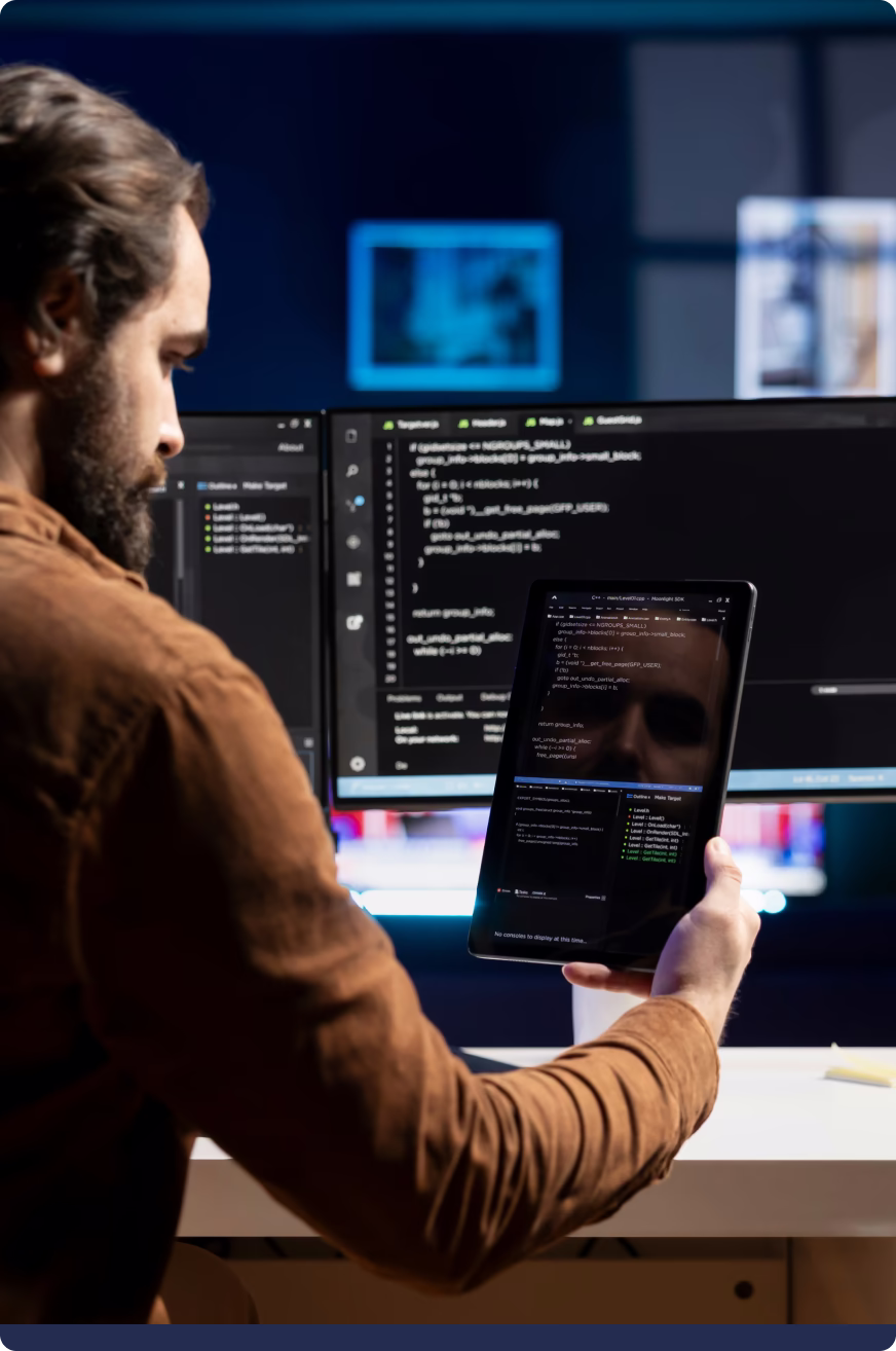 Man holding tablet showing computer code in front of a monitor with software code on screen.