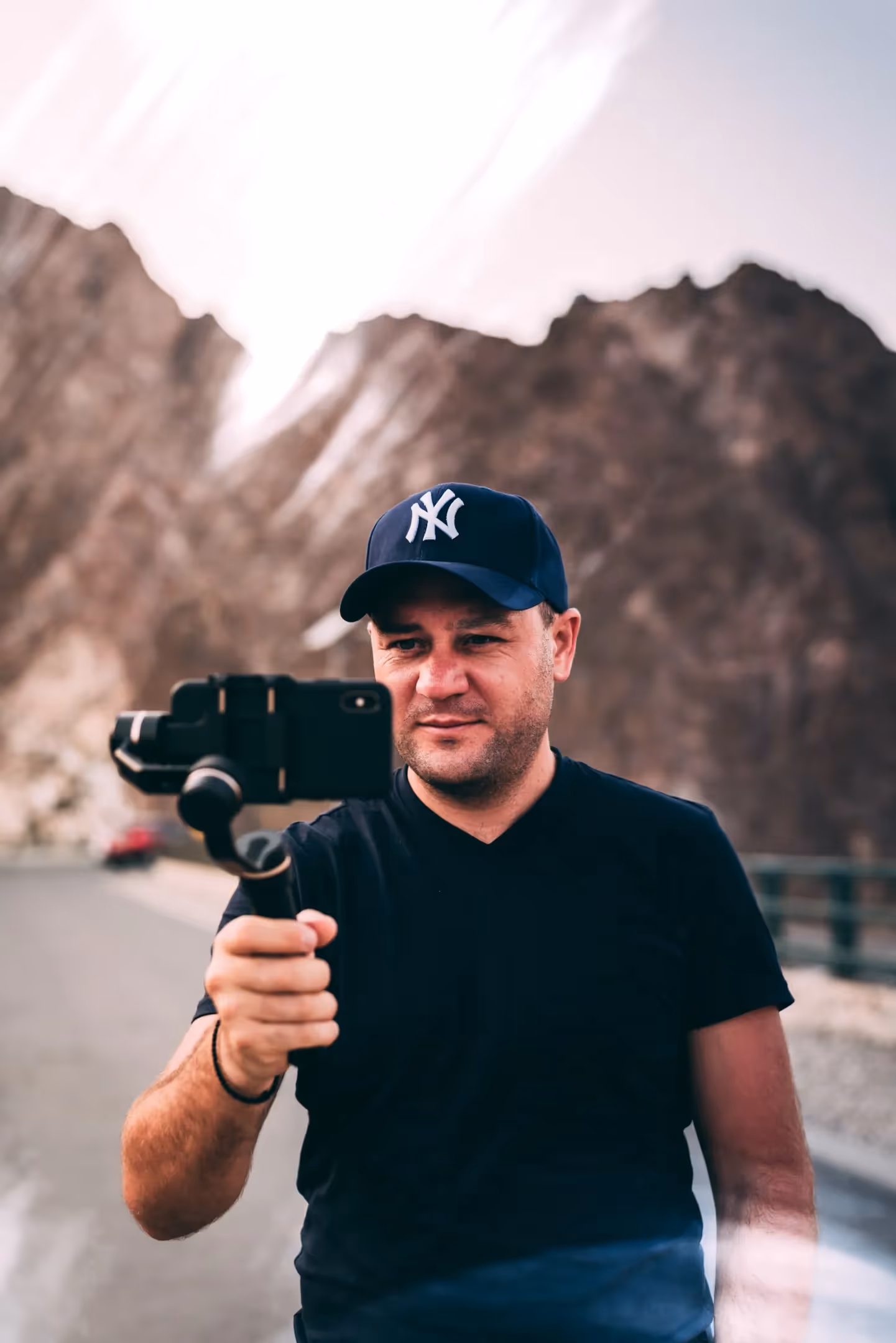 An influencer looking into a handheld tripod with a smartphone attached to it. We can see mountains in the background