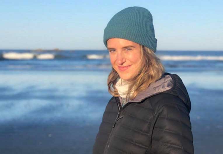 Smiling woman wearing a blue knit beanie and black jacket standing by the ocean with waves in the background.