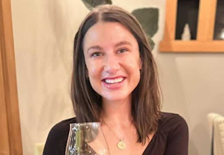 Smiling woman with shoulder-length brown hair holding a wine glass indoors.