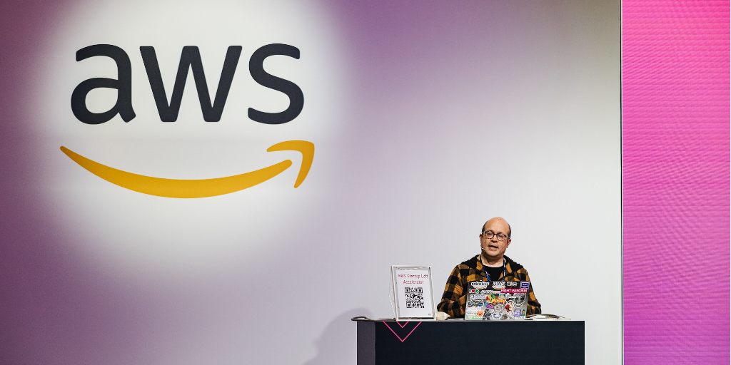 Photo. A man sits behind a small desk in front of a large screen that features the AWS logo on it. On his desk is a QR cod sign and a laptop computer covered in company logo stickers