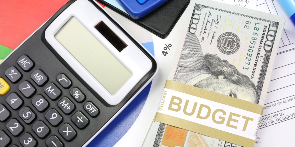 A calculator and stack of 100 dollar bills sit on a messy desk. The stack of bills is wrapped with a paper label that reads "BUDGET"