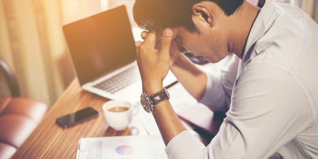 Man sits at desk in front of coffee, phone and computer with his head in his hands.