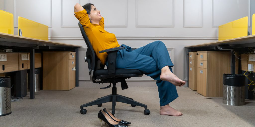 Woman leans back in office chair with arms overhead and shoes off.