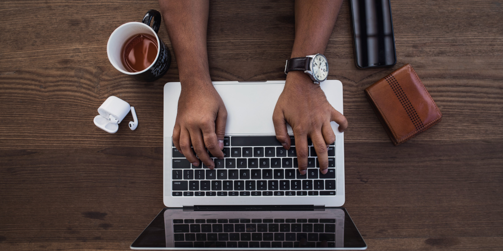 An overhead photo of a person typing on a laptop at a wooden desk. On the desk is also a cup of coffee, notebook, phone and set of Airpods.