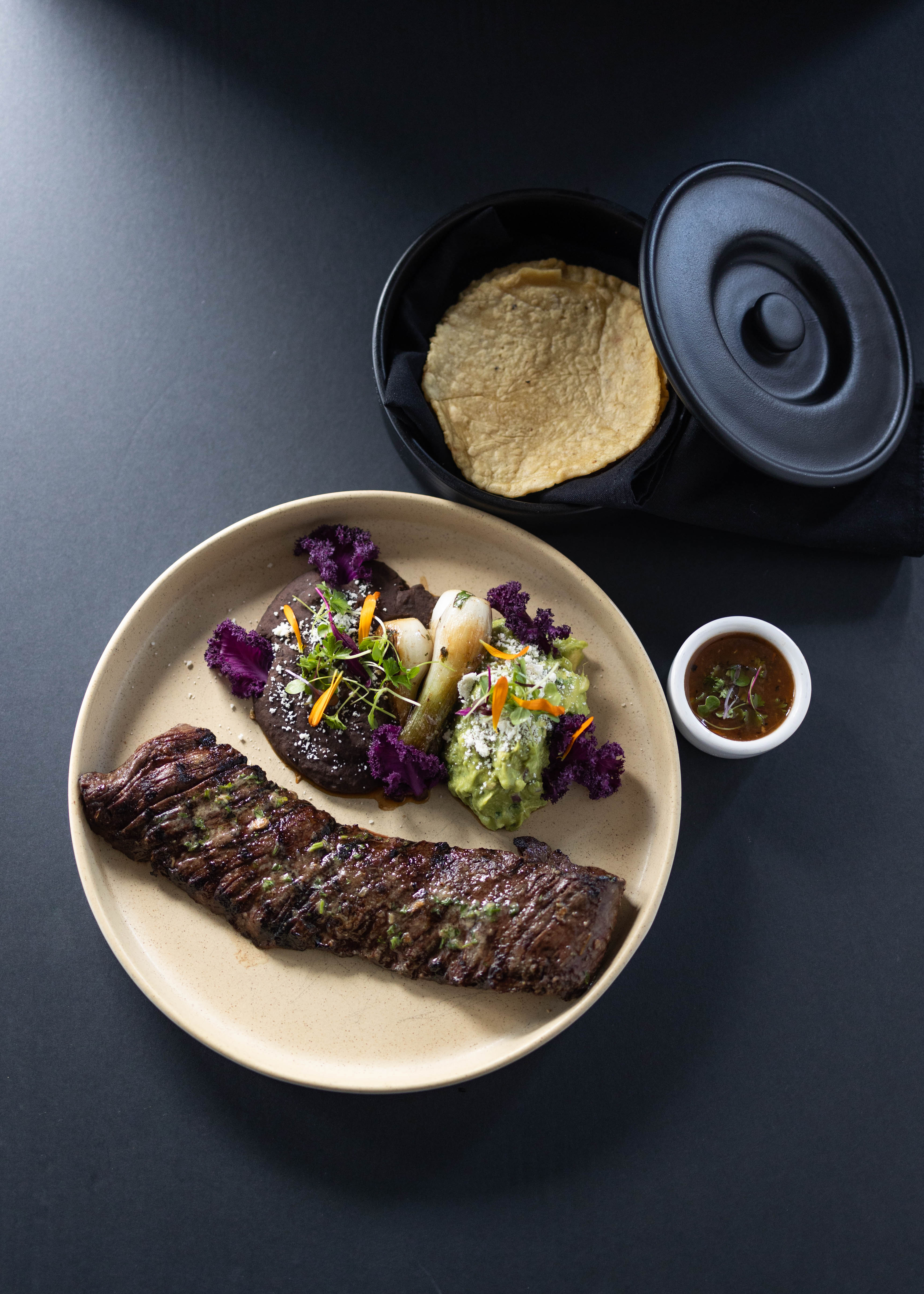 Our popular skirt steak plate. Grilled skirt steak accompanied with refried black beans, guacamole and grilled onions with a side of salsa, and side of handmade corn tortillas