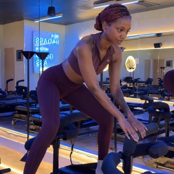 Woman in purple workout clothes stretching forward on gym equipment in a mirrored studio.