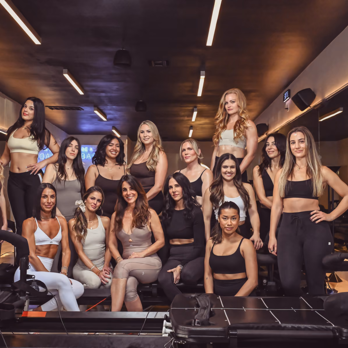 Group of fit women posing in a gym studio wearing athletic wear with exercise equipment visible.