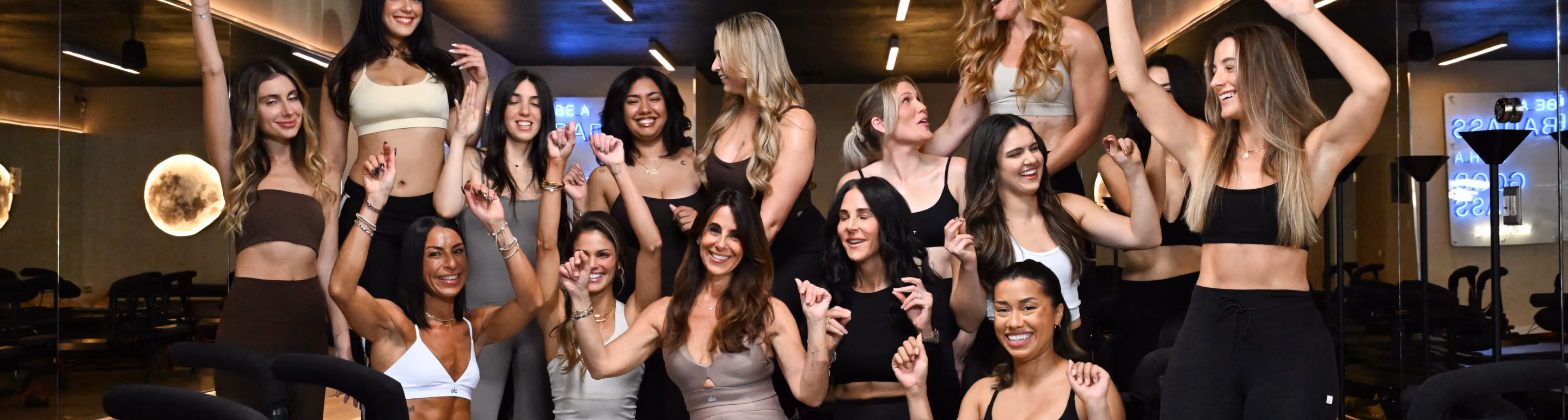 Group of diverse women in fitness attire smiling and raising arms in a gym or studio setting.