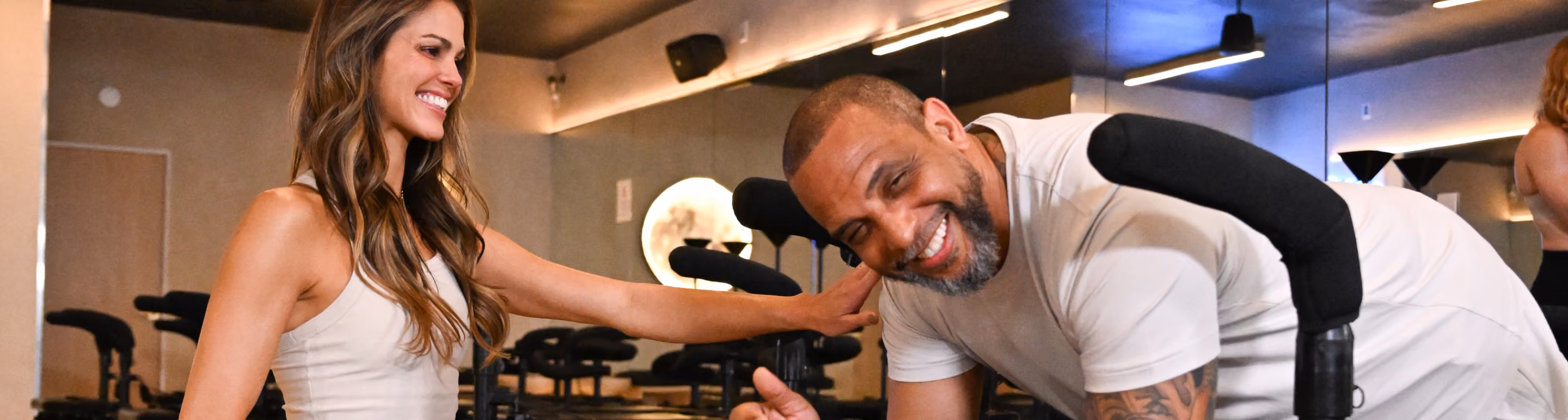 Smiling woman assists man in light workout in a fitness studio with equipment in the background.