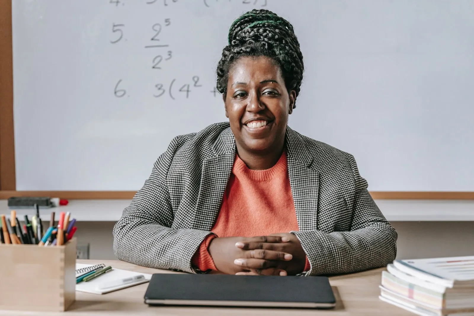 A math teacher is seated in front of a whiteboard with her hands together on the desk.