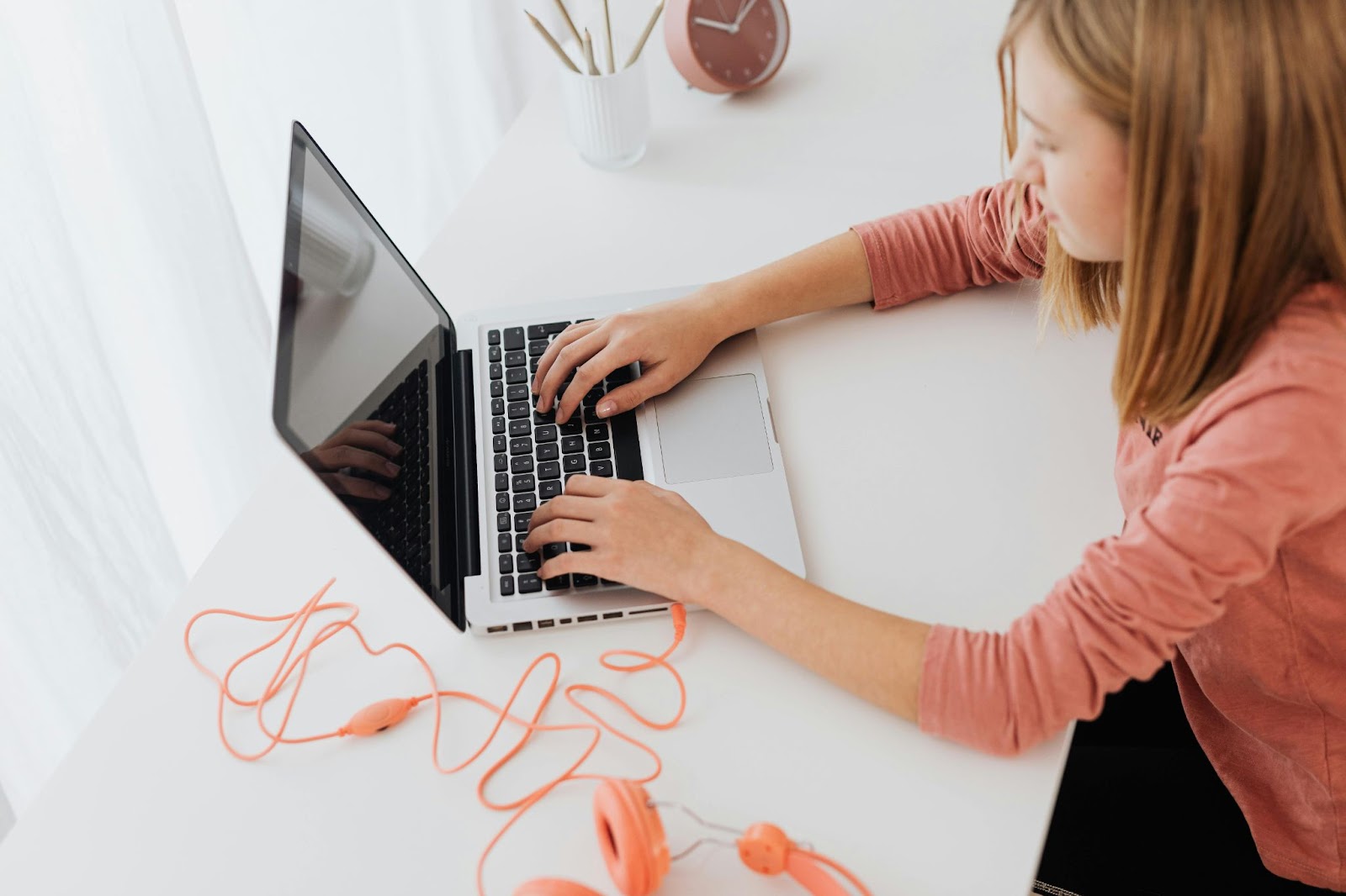 A young girl types on her laptop. Her bright orange headphones are stretched out beside the computer.y