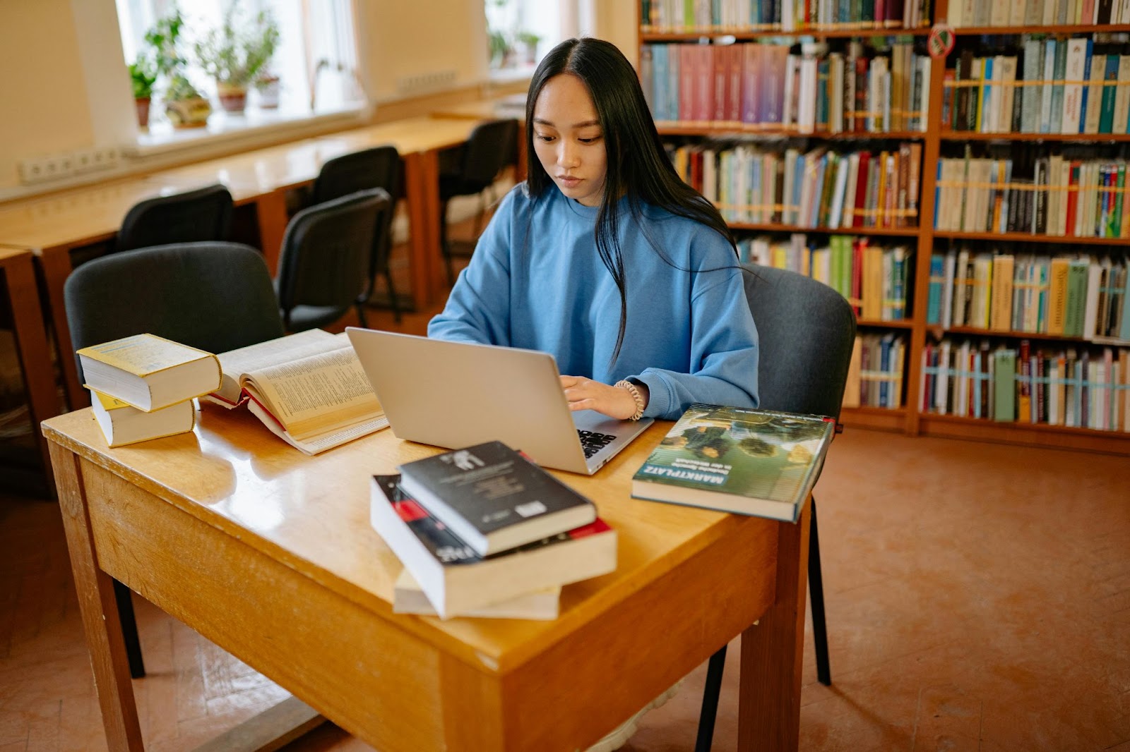 A student works on her laptop at the library, surrounded by stacks of books.