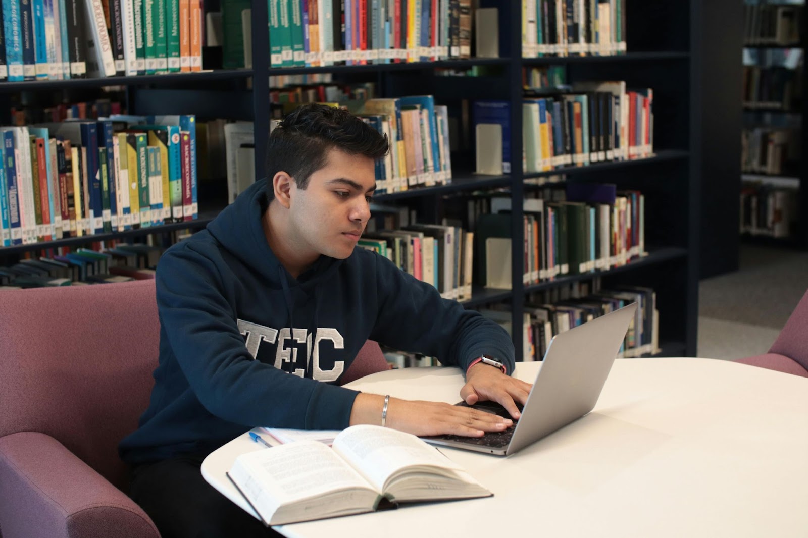 A student works on his laptop at the library with a book open on the table next to him.