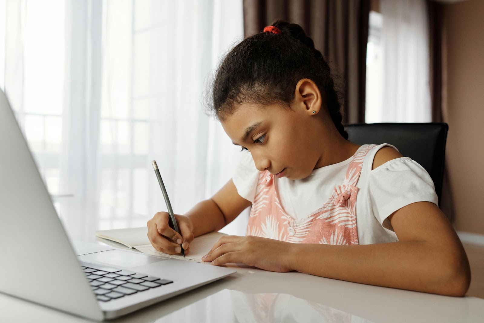 A young girl writes in her notebook with a laptop open in front of her.