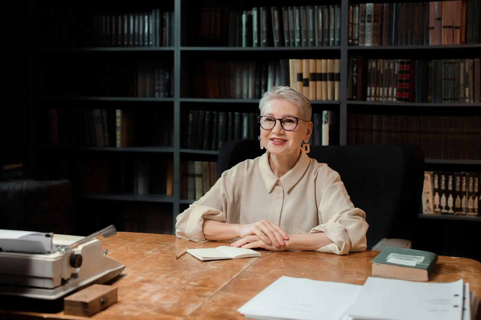 A smartly dressed female administrator sits at her desk with her hands crossed. Behind her is a large library of dark-covered books.