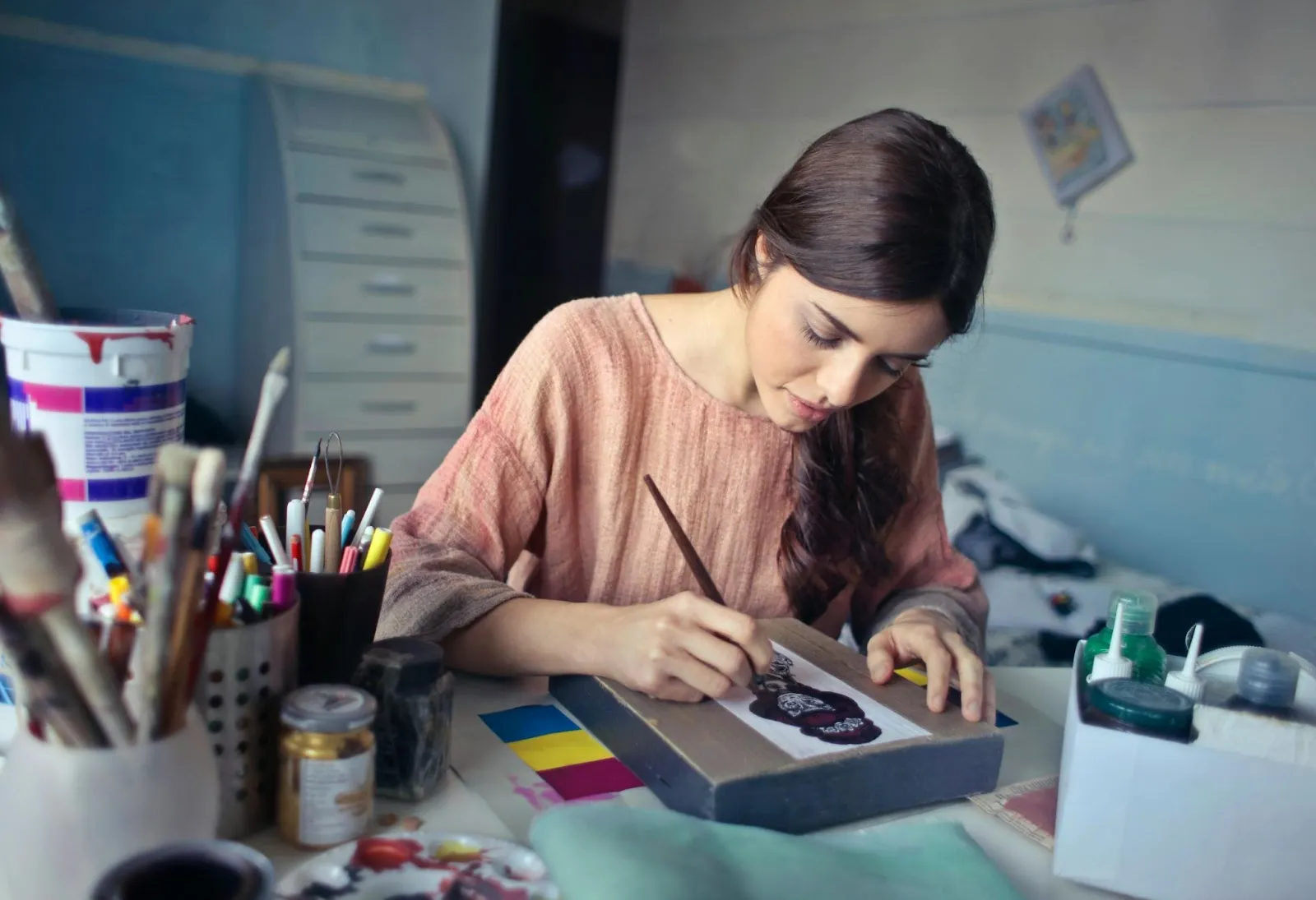 A woman paints at her crowded crafting desk.