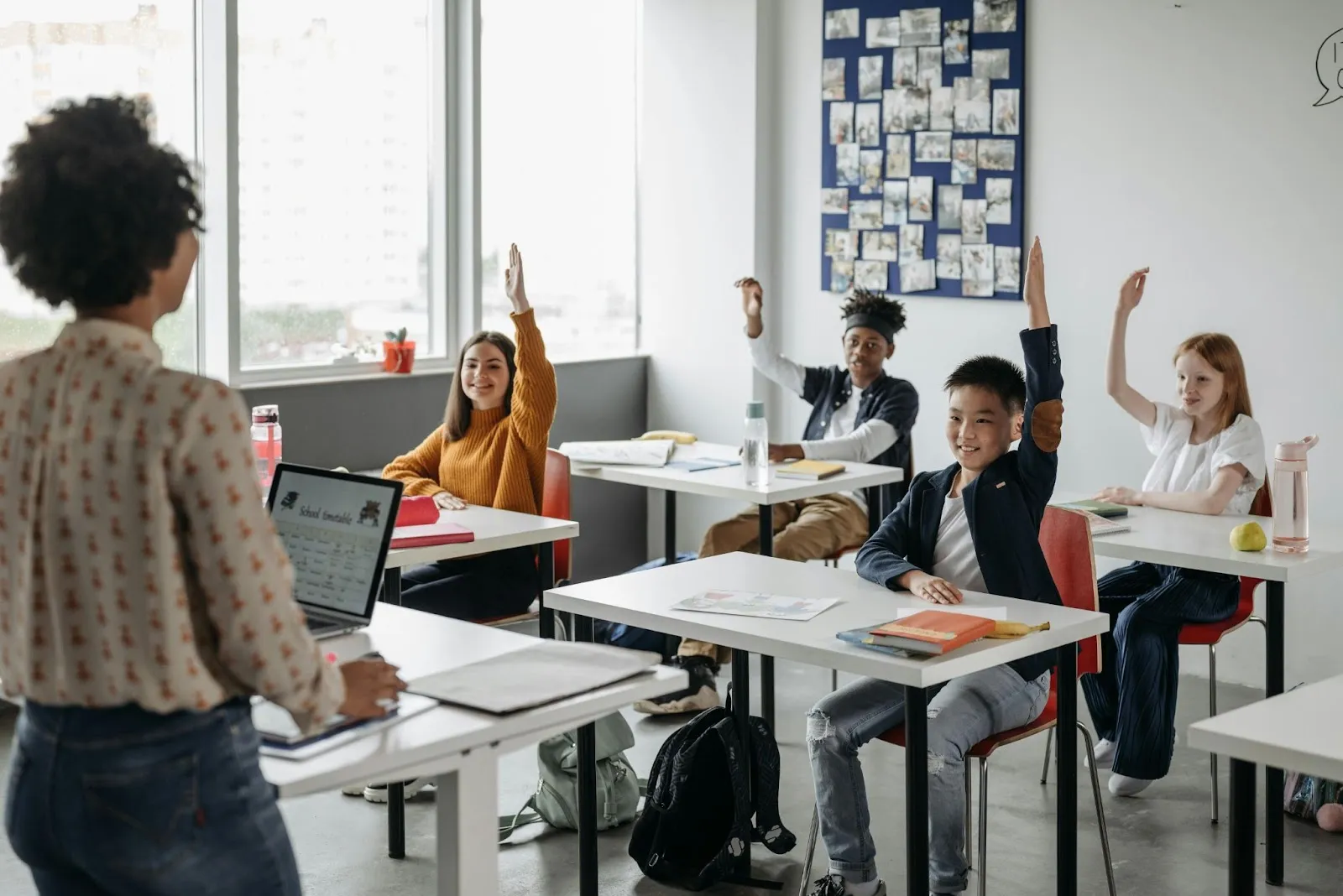 A teacher stands in front of a small classroom of students politely raising their hands.