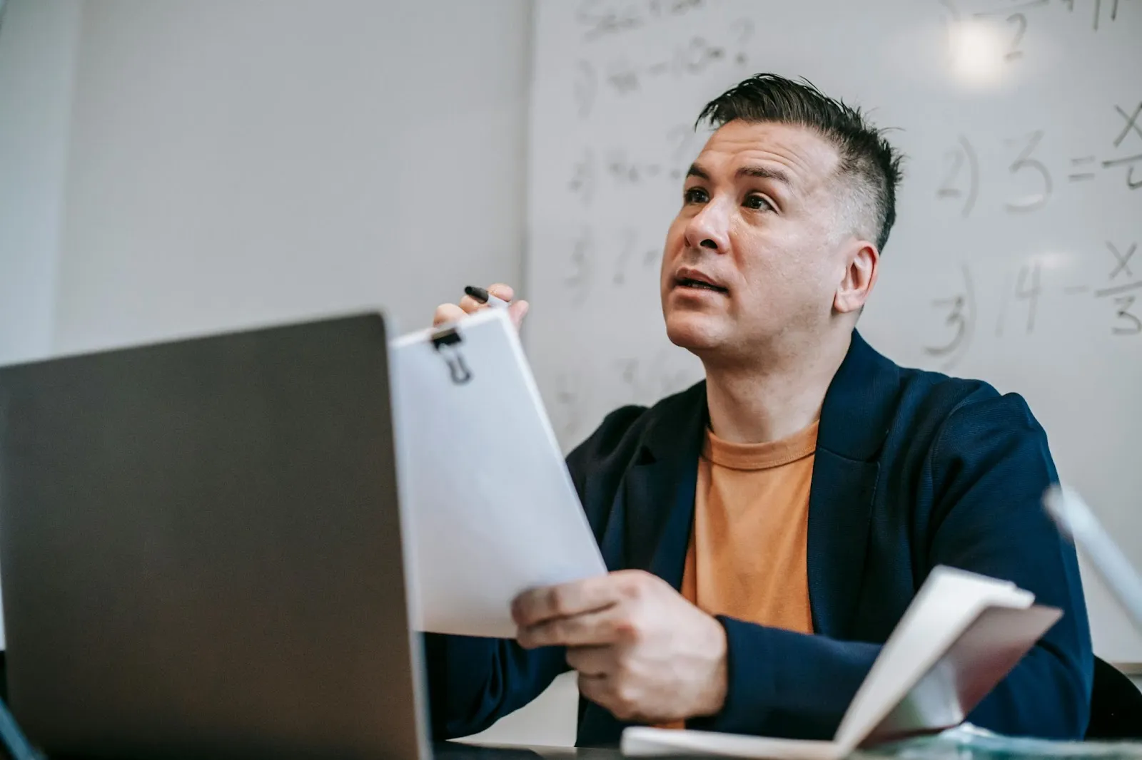 A teacher discusses a stack of paperwork over the top of his laptop screen to an invisible conversant.