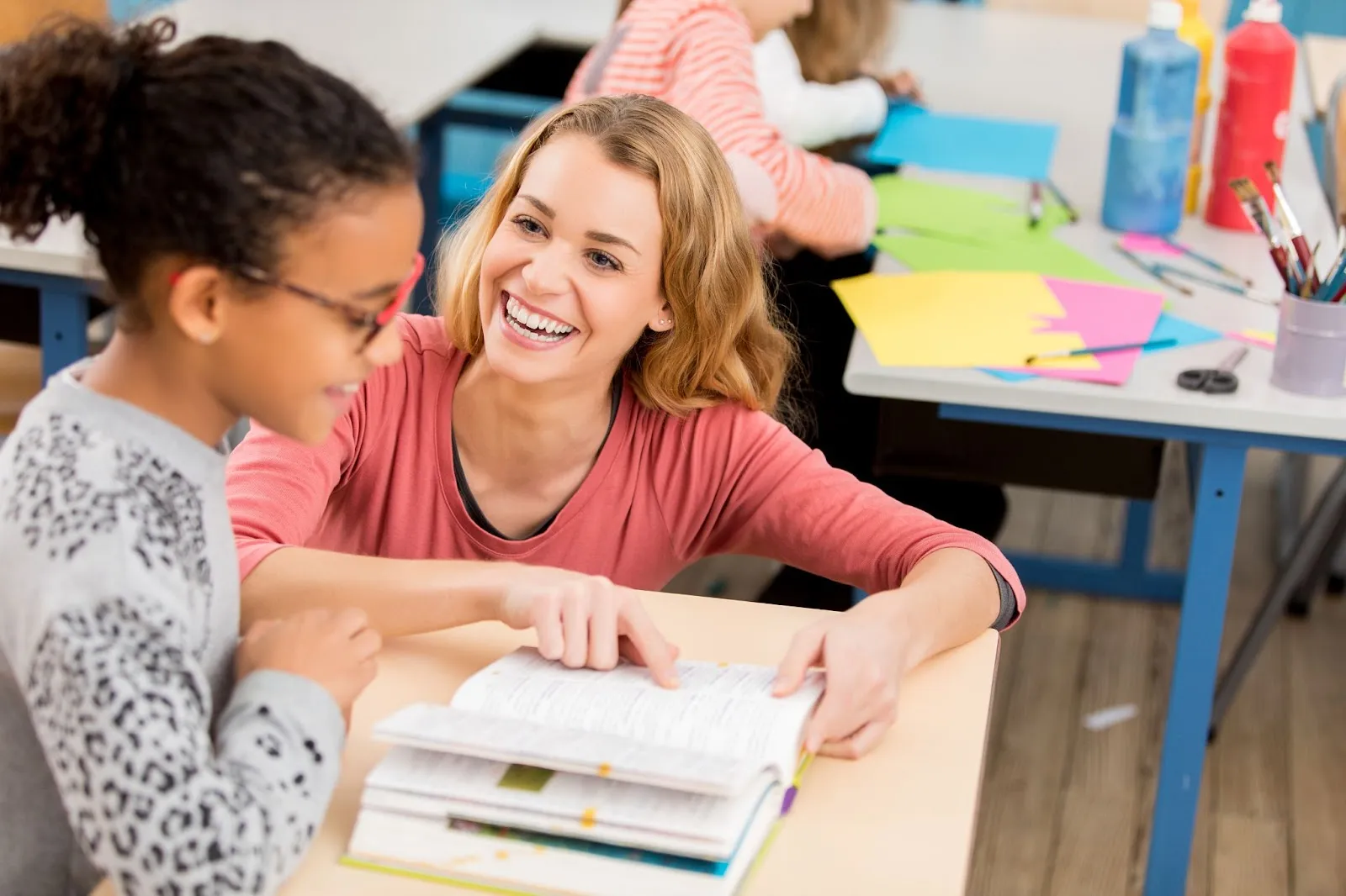 A teacher kneels next to her student's desk to help with her textbook.