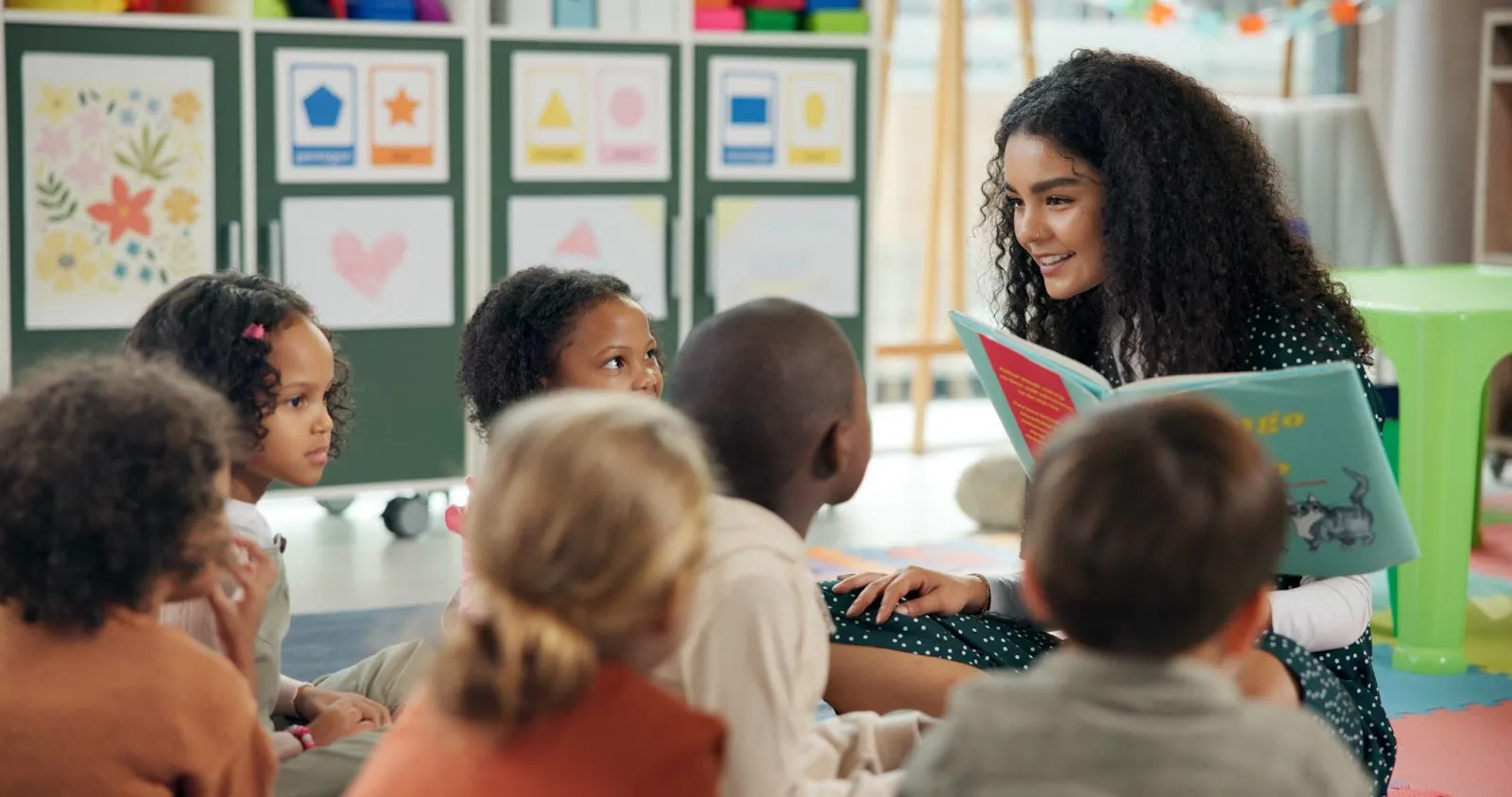 A young teacher reads a book to a small group of young students. She is engaged and surrounded by pops of colorful decoration.