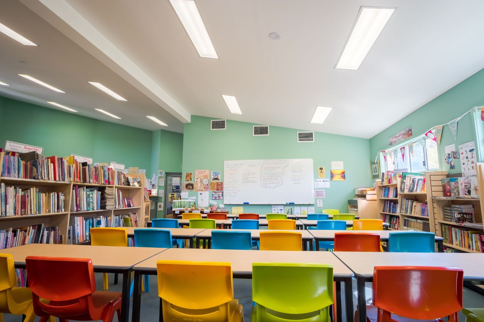 The classroom is surrounded on either side by full shelves of books. Rows of tables face the front of the room, chairs are colorful and the walls are painted green.