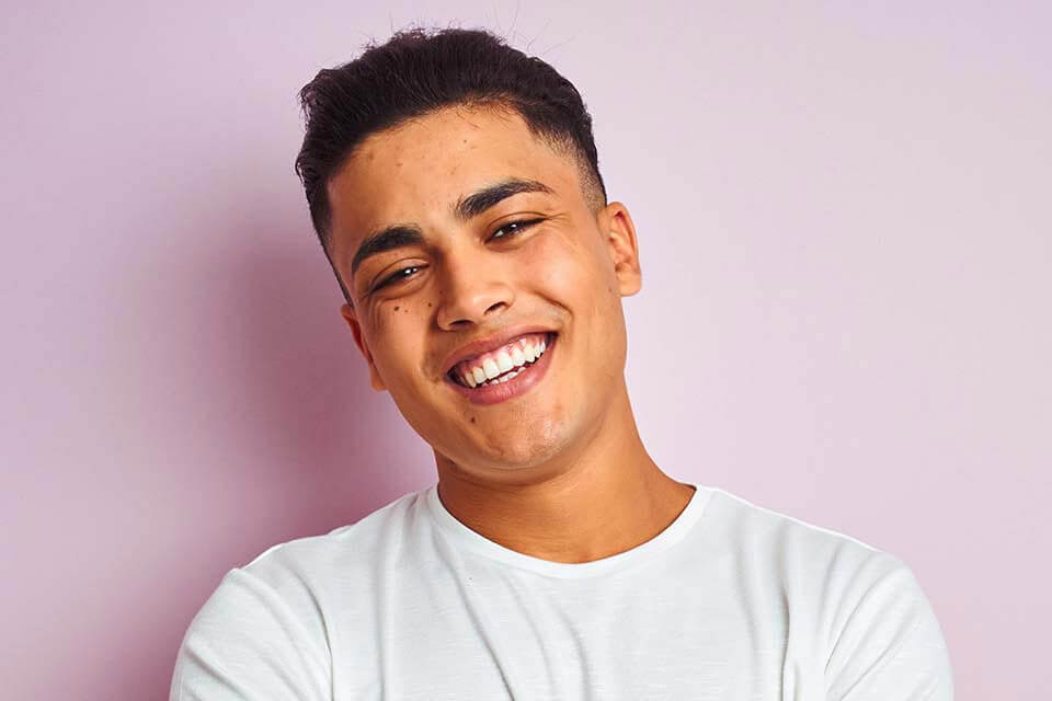 Smiling young man with short dark hair wearing a white t-shirt against a light purple background.