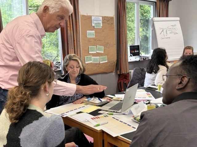 Sam Boering leans over a table to point out a step in a results chain while participants look on, flip charts of a theory of change and indicators visible behind him.