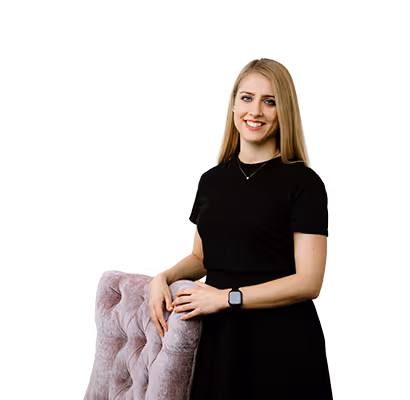 Smiling woman with long blonde hair wearing a black dress and smartwatch, standing with her hand on a mauve tufted chair.