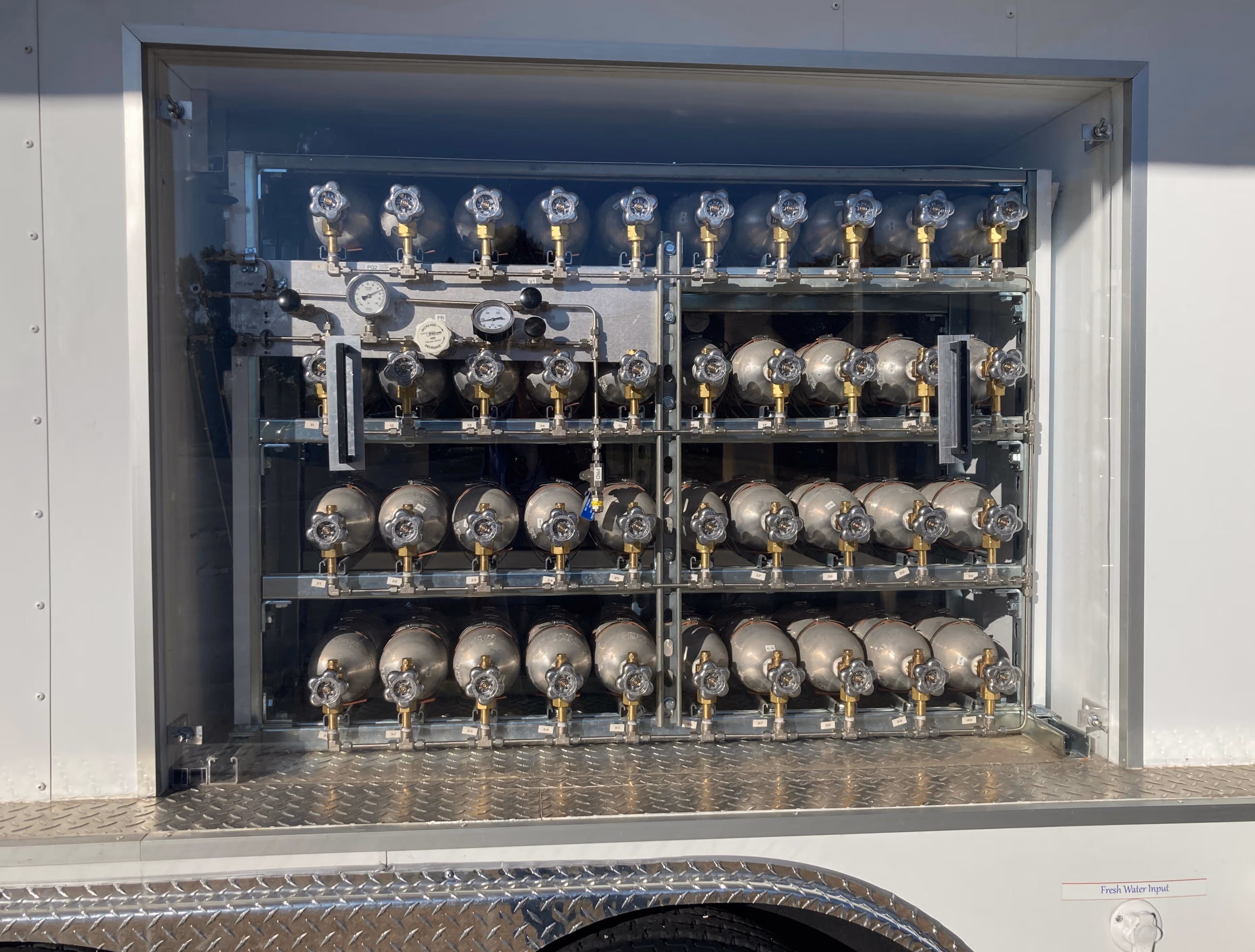 Compartment with multiple arranged metal gas cylinders and pressure gauges behind glass on a vehicle.