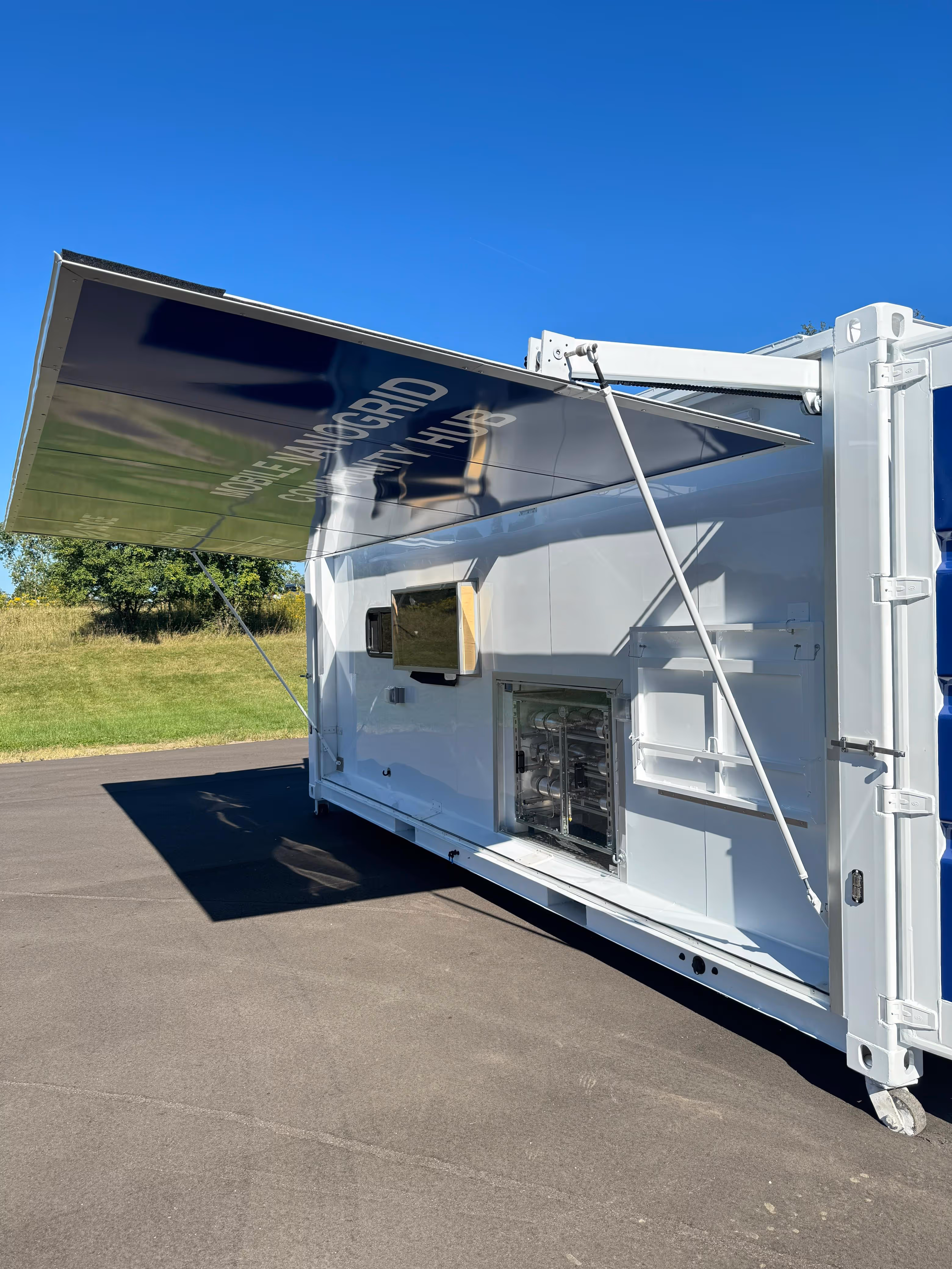 White mobile nanogrid community hub container with an opened side panel revealing equipment inside, parked on an asphalt surface under clear blue sky.