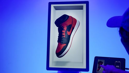 Hololuminescent™ Display (HLD) showing a red and black Nike sneaker floating in a white hologram embed with a person interacting on a tablet nearby.