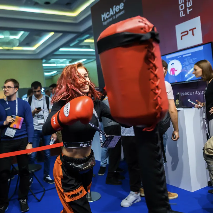 A young woman in boxing gloves punches a large red boxing mitt at a crowded indoor event.