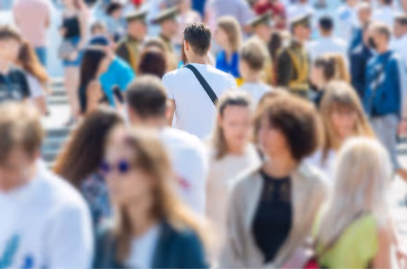 Crowd of people walking outdoors, with one person in a white shirt and black shoulder bag in focus from behind.