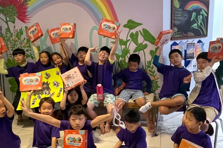 Group of children in purple shirts holding up orange boxes and a yellow poster in a colorful classroom with a rainbow and plant mural.