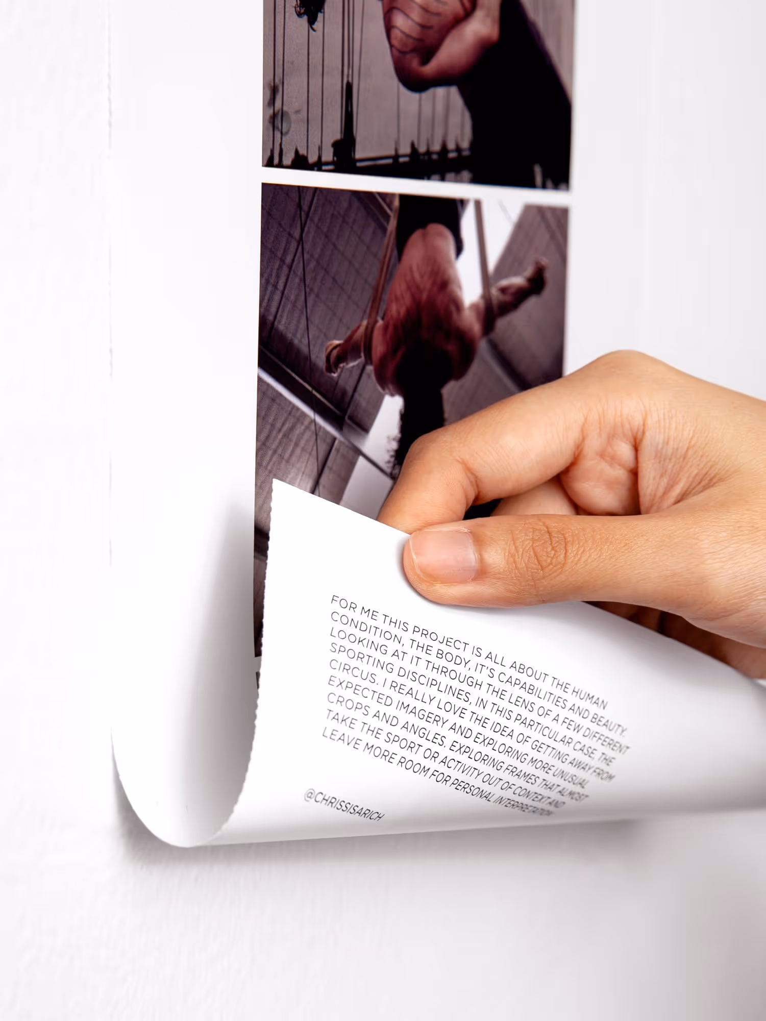 Hand peeling back a corner of a printed poster showing a man performing aerial acrobatics on rings inside an industrial setting.