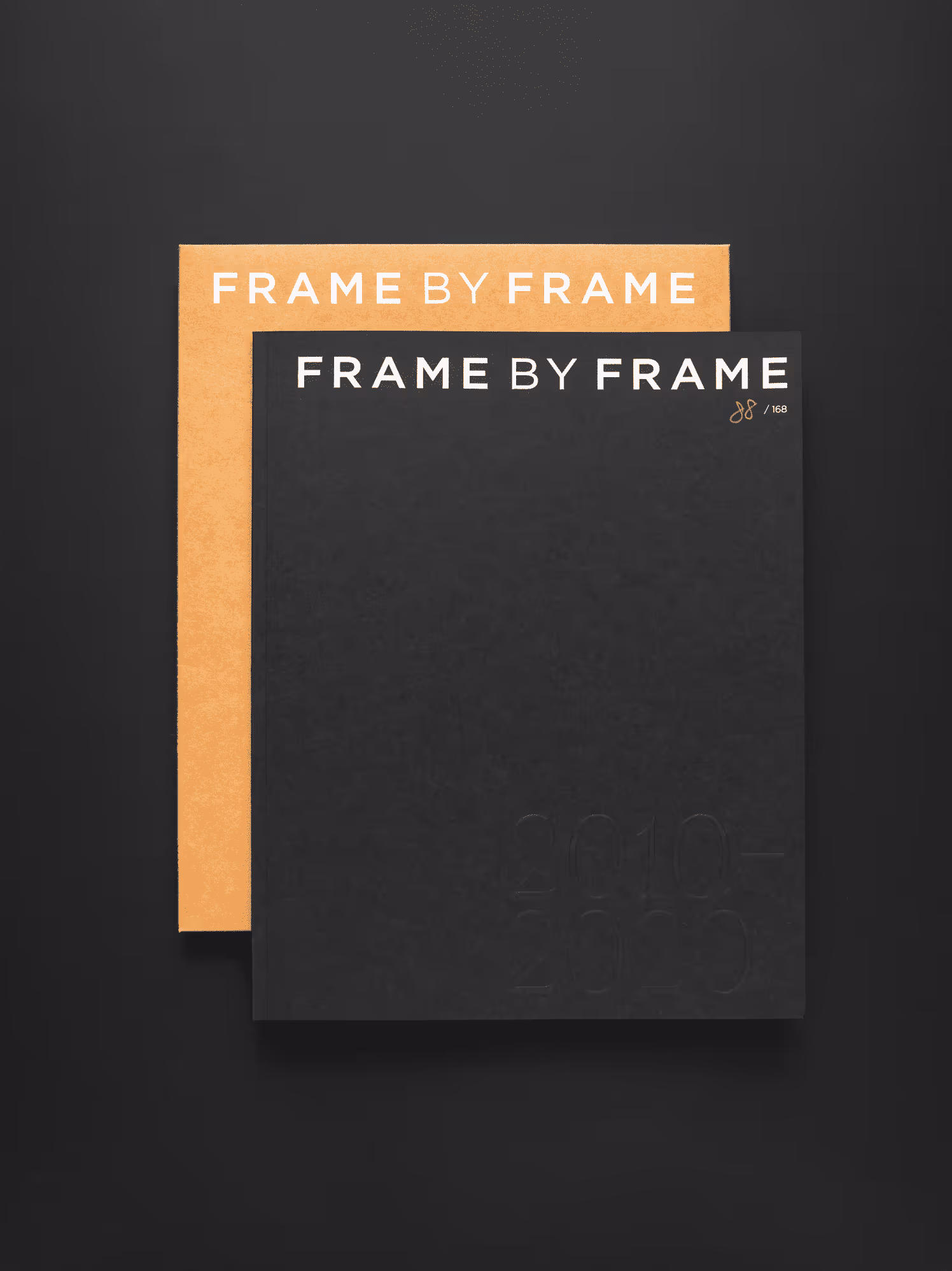 Two stacked books titled 'FRAME BY FRAME' with one in black and the other in light brown on a black surface.