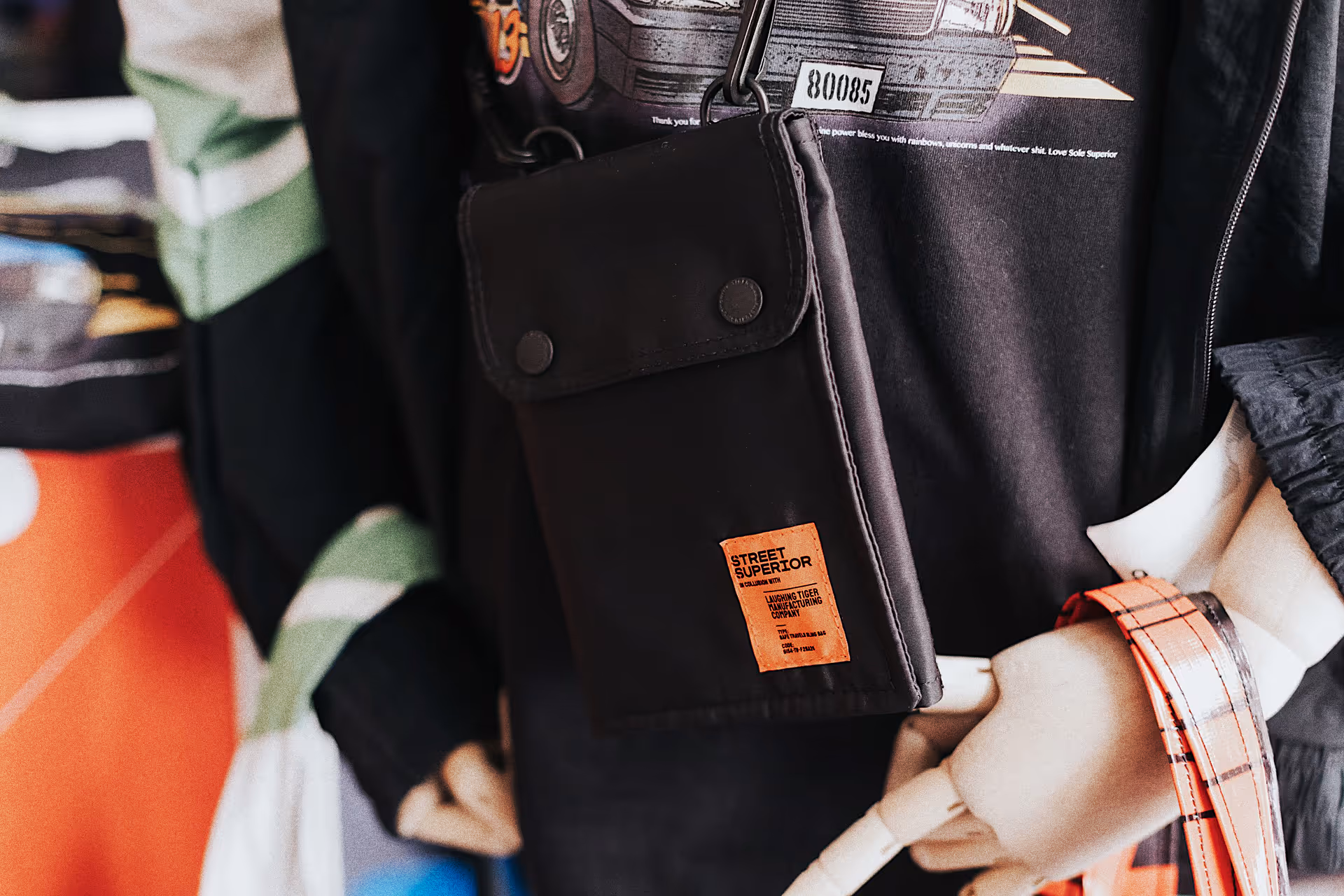 Black crossbody bag with orange Street Superior label worn over a black graphic t-shirt with a mannequin hand holding the strap.