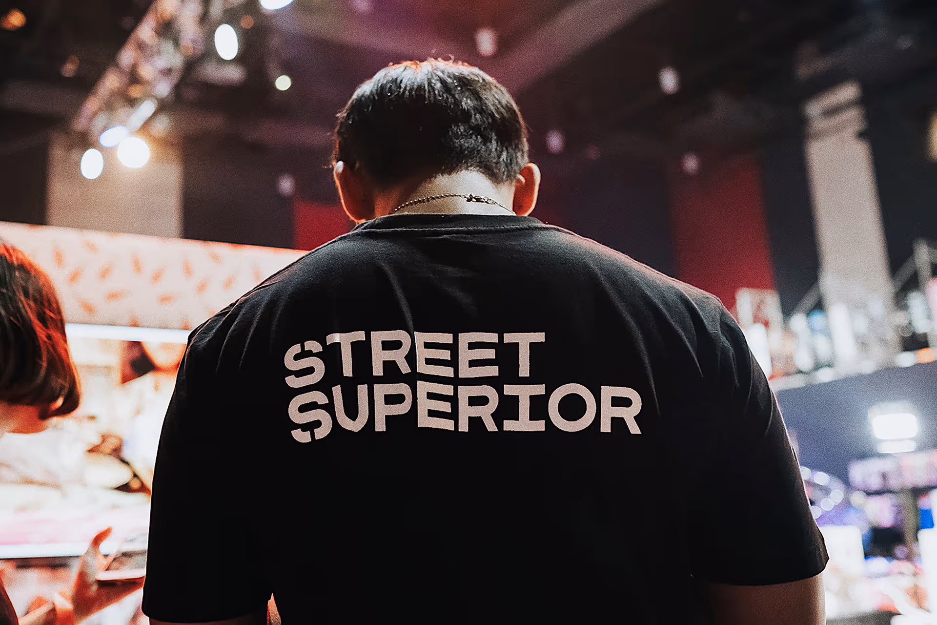 Person facing away wearing a black shirt with the words 'STREET SUPERIOR' in white letters on the back in an indoor event setting.