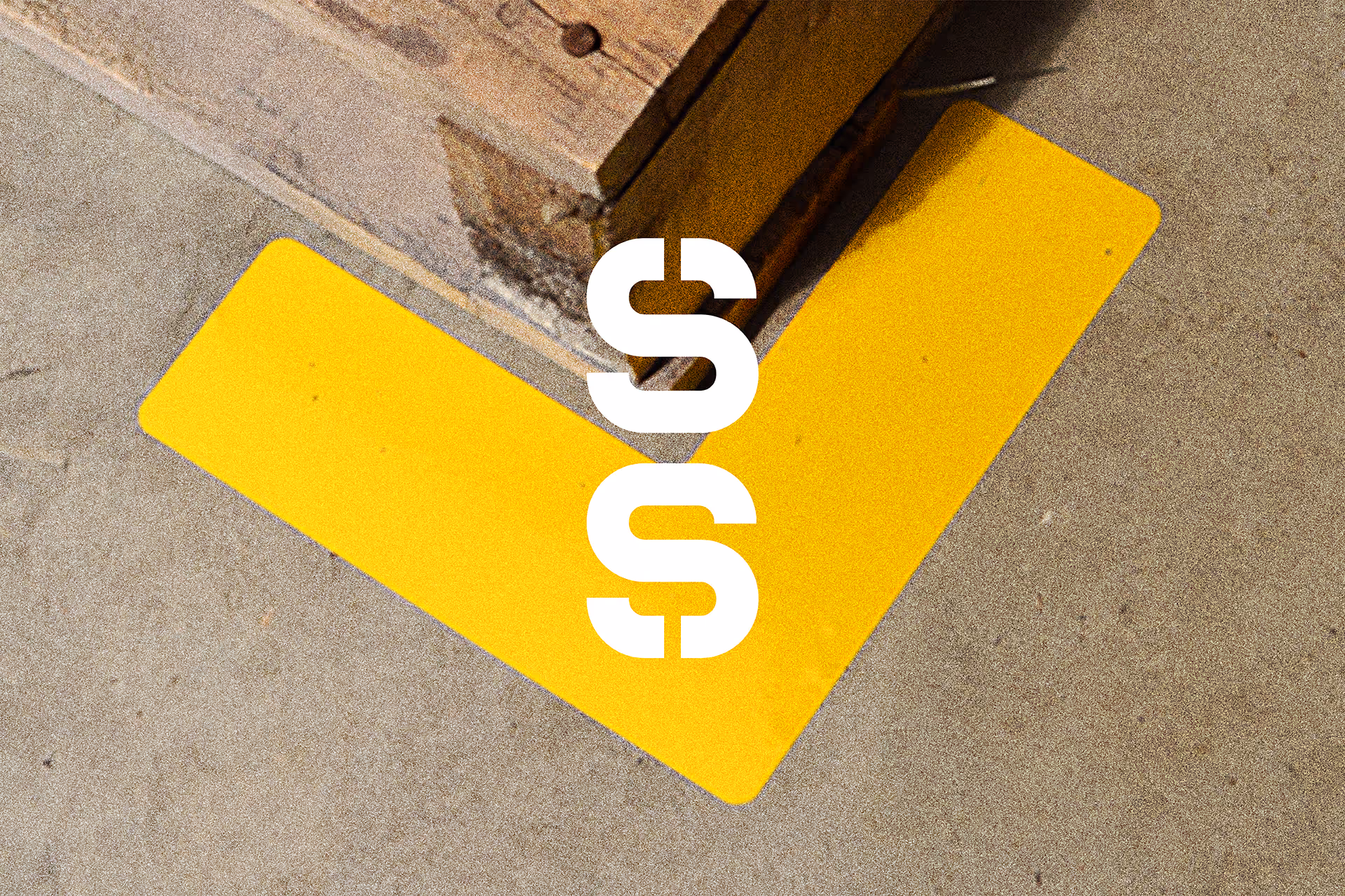 Yellow L-shaped tape on concrete floor next to a wooden structure corner with white stylized double 'S' letters centered above it.