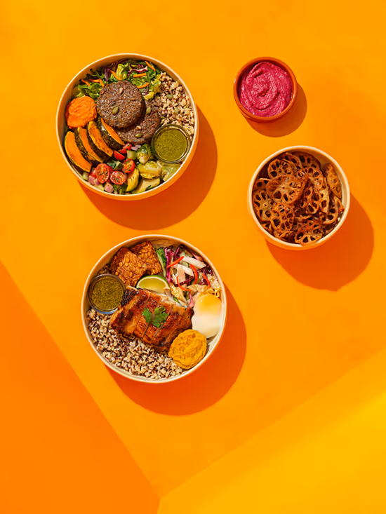Top view of two bowls with rice, grilled vegetables, tempeh, and sauces, accompanied by a bowl of lotus root chips and a small bowl of beet dipping sauce on an orange background.