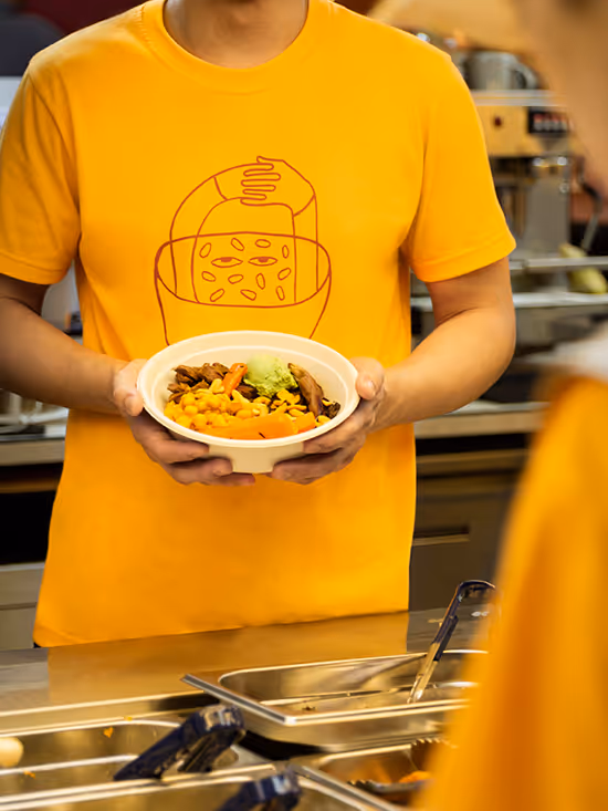 Person in yellow shirt holding a bowl of food with corn, carrots, and other vegetables at a food counter.