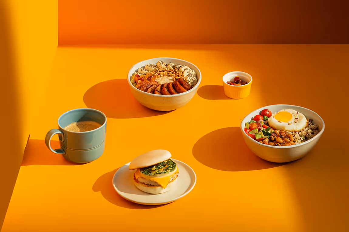 Breakfast spread on an orange surface including a blue cup of coffee, a sandwich with cheese and egg on a white plate, two bowls of mixed grains and vegetables topped with sausage and a sunny-side-up egg, and a small dish of berries.