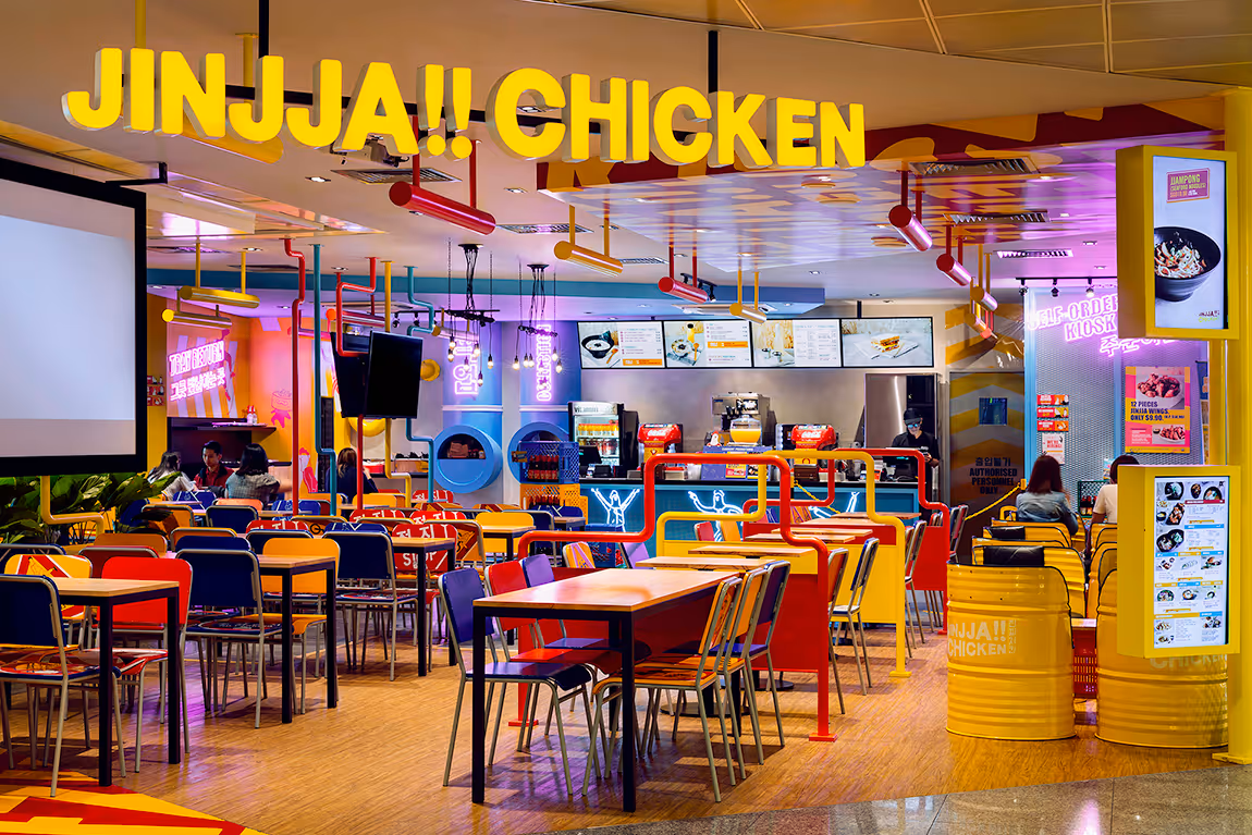 Interior of Jinjja Chicken restaurant with colorful chairs, wooden tables, neon signs, and digital menu screens at the counter.