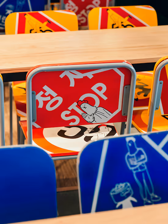 Colorful chairs with graphic designs including a red stop sign with a praying chicken and other artistic images, arranged around wooden tables.