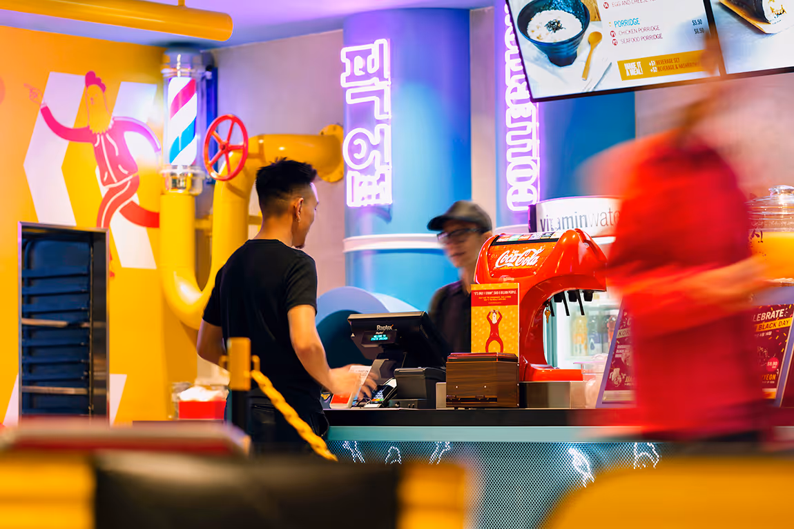 Man in black shirt standing at a colorful restaurant counter with a Coca-Cola soda dispenser and neon signs in the background.