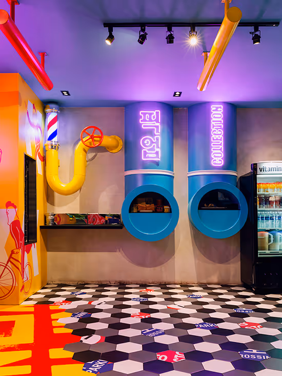 Colorful indoor space with two large blue circular collection tubes labeled in neon text, a yellow pipe structure with a red valve, and a geometric patterned floor.