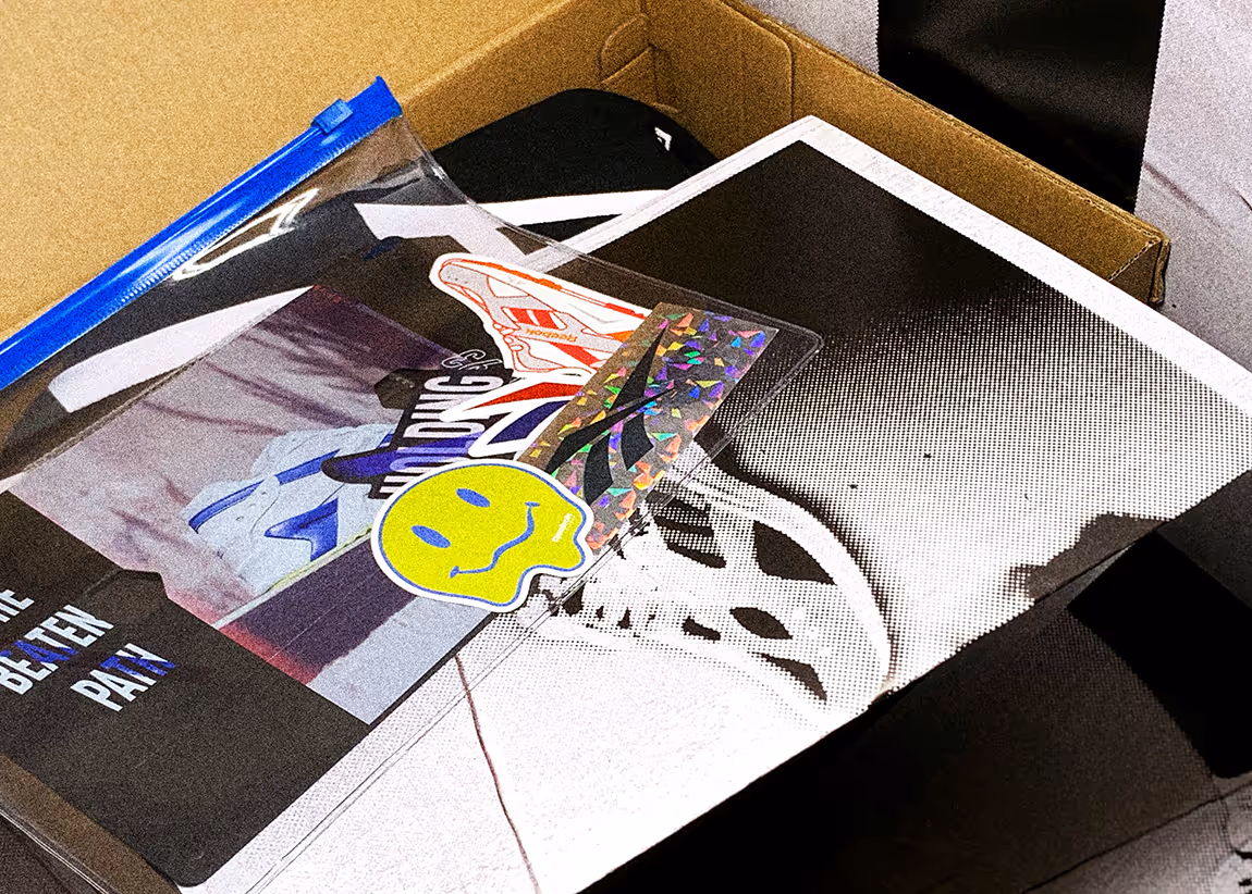 Close-up of a partially open cardboard box containing a book with a black and white halftone shoe design and a clear zipper pouch with colorful stickers including a melting yellow smiley face and shoe-themed graphics.